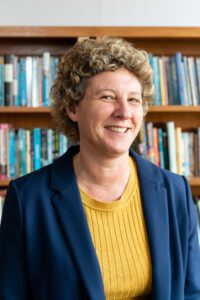 Teacher in yellow top and blue blazer in front of book case